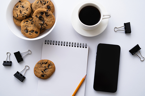 top view of coffee, cookies and stationery near smartphone with blank screen on white