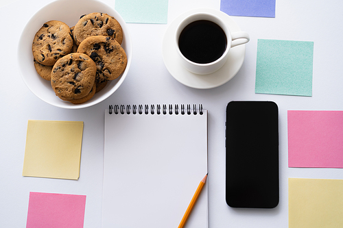 top view of cup of coffee, cookies and stationery near smartphone with blank screen on white