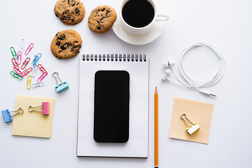 top view of cup of coffee, biscuits and smartphone with blank screen near stationery on white