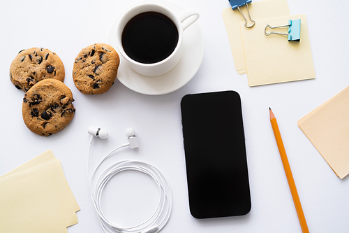 top view of cup of coffee and chocolate chip cookies near stationery and smartphone on white