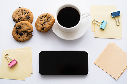 top view of cup of coffee and chocolate chip biscuits near stationery and smartphone on white
