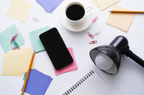 top view of cup of coffee near stationery, lamp and smartphone with blank screen on white