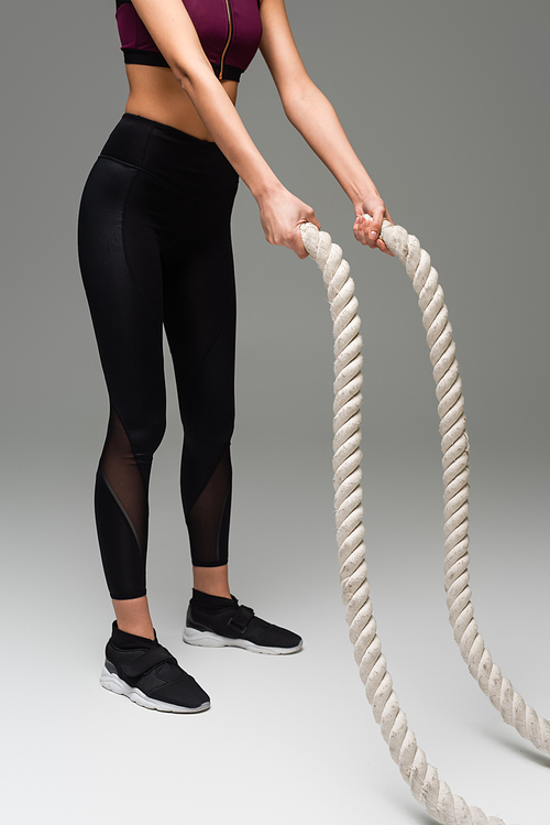 partial view of sportswoman in black leggings and sneakers training with battle ropes on grey background