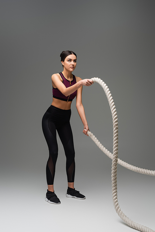 full length view of sportive woman exercising with battle ropes on grey background