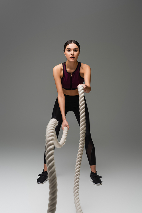 full length view of sportive woman working out with battle ropes on grey background