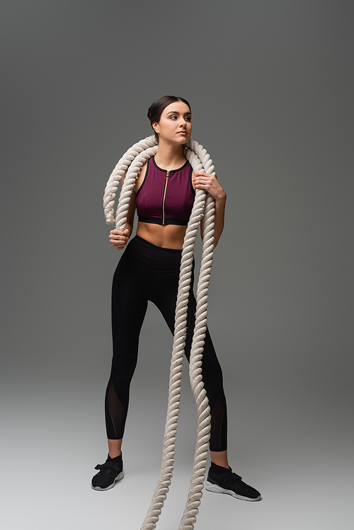 full length view of young sportswoman holding battle ropes on grey background