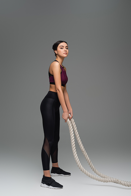 young sportswoman  while standing with battle ropes on grey background