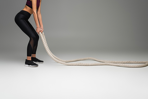 cropped view of sportswoman in black leggings exercising with battle ropes on grey background