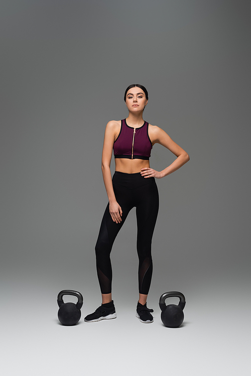 confident sportswoman standing with hand on hip near kettlebells on grey background