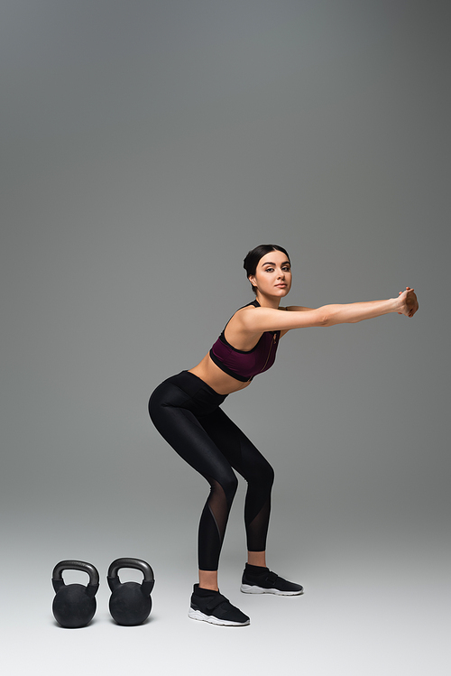 young sportswoman doing sit ups near kettlebells on grey background