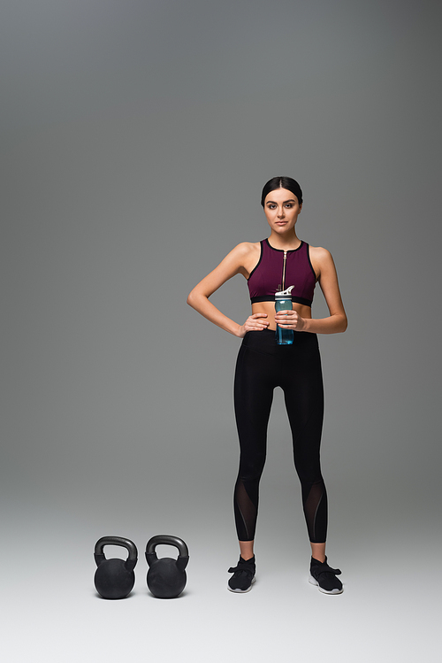 confident woman with sports bottle standing near kettlebells on grey background