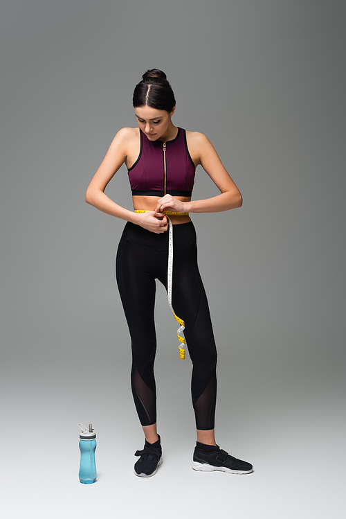 full length view of slim woman in black sportswear measuring waist on grey background