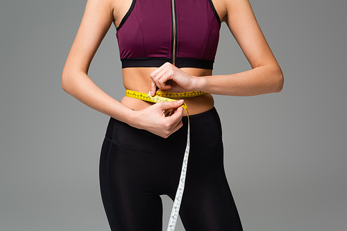 cropped view of woman in black leggings and sports top measuring waist isolated on grey