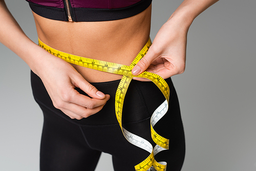 partial view of sportswoman measuring waist isolated on grey