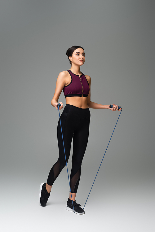 full length view of woman in black sportswear with skipping rope on grey background