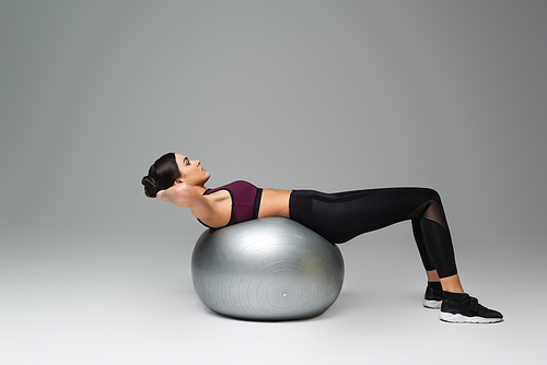 side view of woman in black sportswear training on fitness ball on grey background