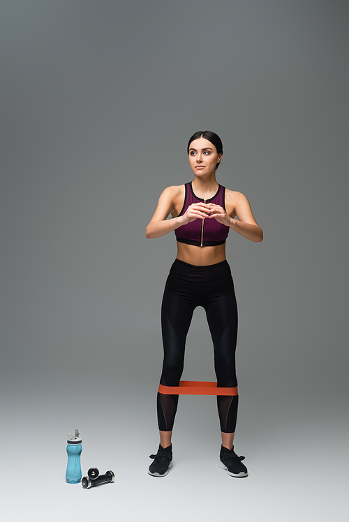 full length view of sportive woman looking away while training with elastics on grey background