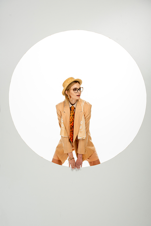 Fashionable girl in beige jacket and straw hat looking away near round hole on white background