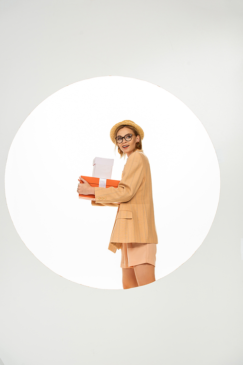 Side view of smiling girl in beige jacket and straw hat holding presents on white background