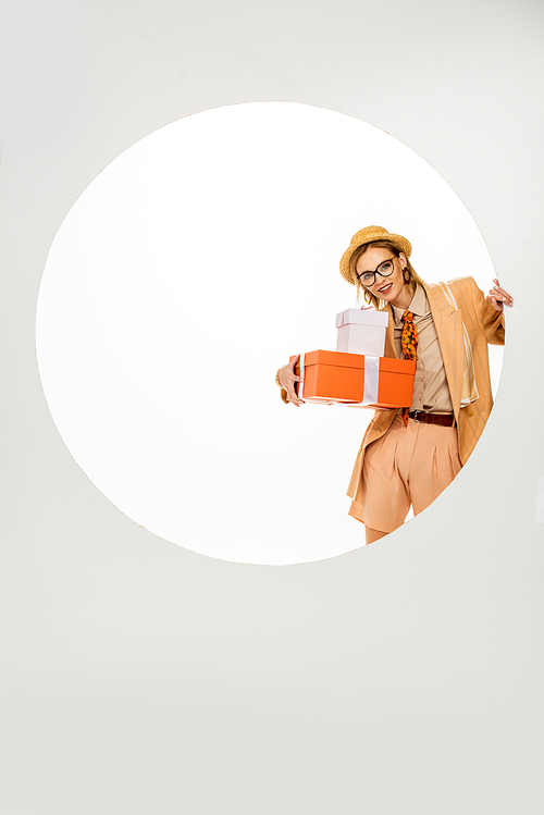 Trendy woman smiling while holding presents near round hole on white background