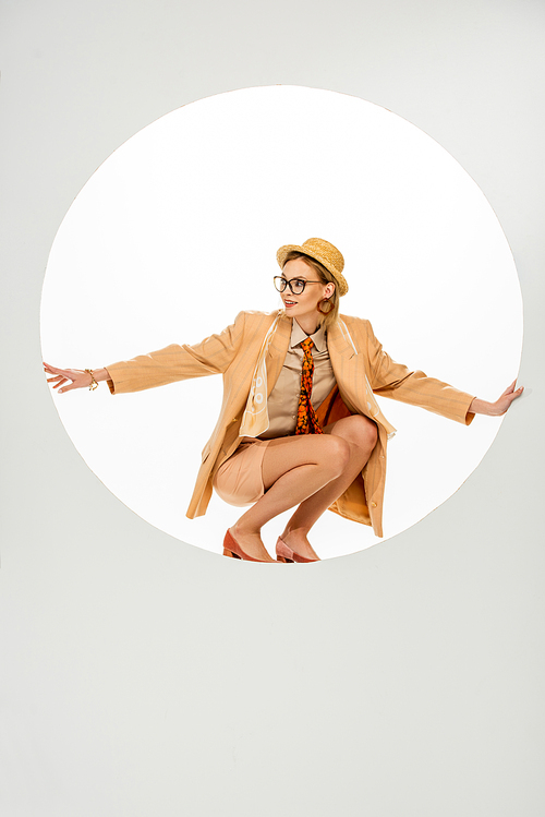 Smiling woman in beige blazer and straw hat standing in round hole on white background