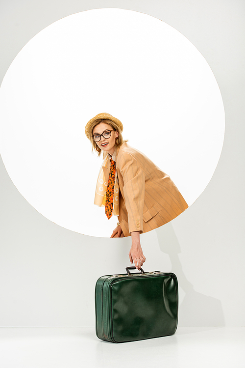 Side view of stylish girl smiling at camera while holding green travel bag near circle on white background