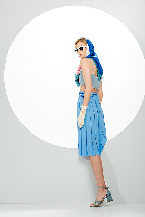 Side view of trendy woman in blue skirt and sunglasses standing near circle on white background