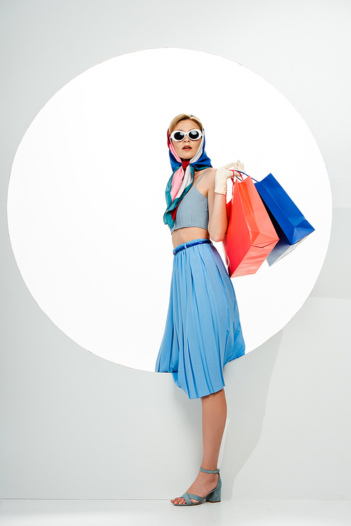 Fashionable woman in sunglasses and headscarf holding blue and red shopping bags near circle on white background
