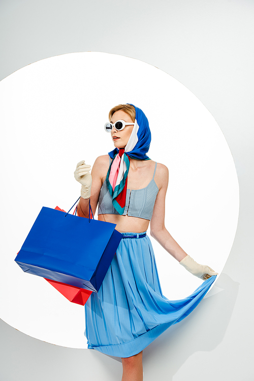 Fashionable girl in gloves and sunglasses holding blue and red shopping bags on white background