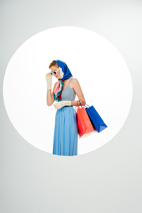 Stylish woman smiling while adjusting sunglasses and holding colorful shopping bags near circle on white background