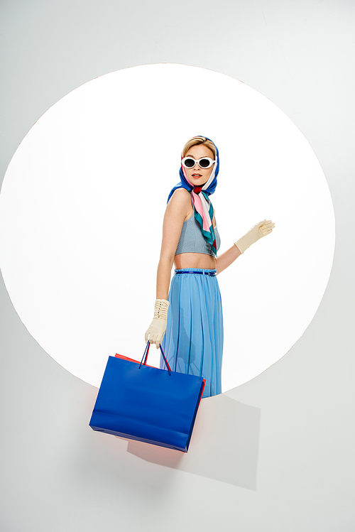 Fashionable girl in headscarf and sunglasses holding shopping bags near circle on white background