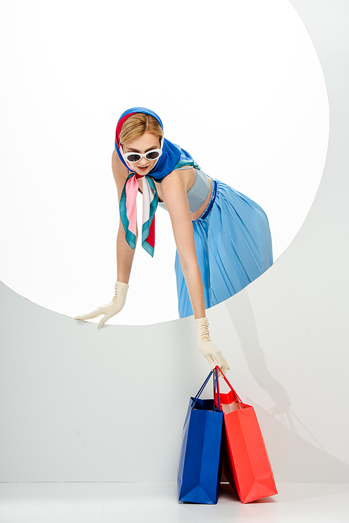 Trendy woman in headscarf and sunglasses putting red and blue shopping bags near circle on white background