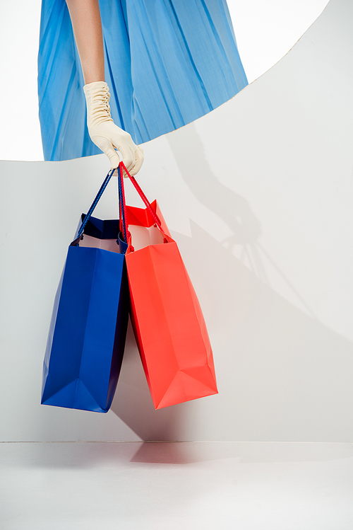 Cropped view of stylish woman in glove holding red and blue shopping bags near round hole on white background