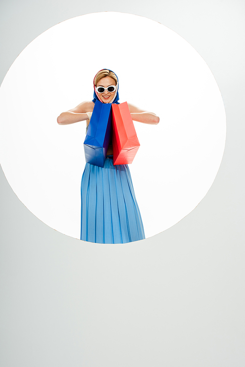 Cheerful woman in sunglasses and headscarf holding red and blue shopping bags near circle on white background