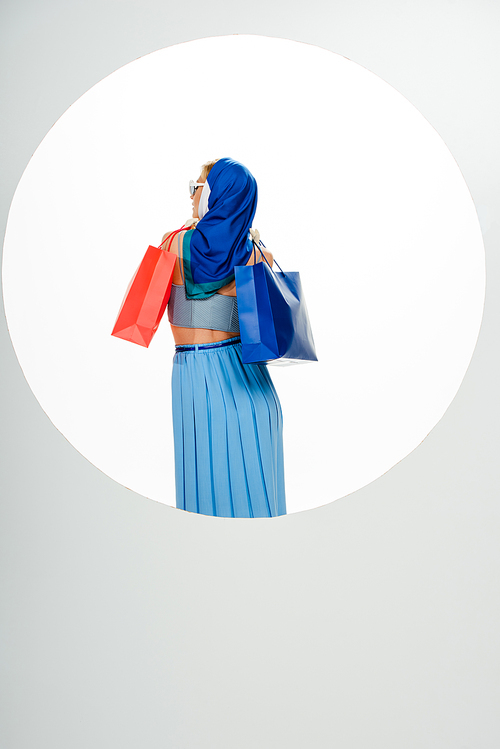 Back view of stylish woman in headscarf holding red and blue shopping bags near circle on white background