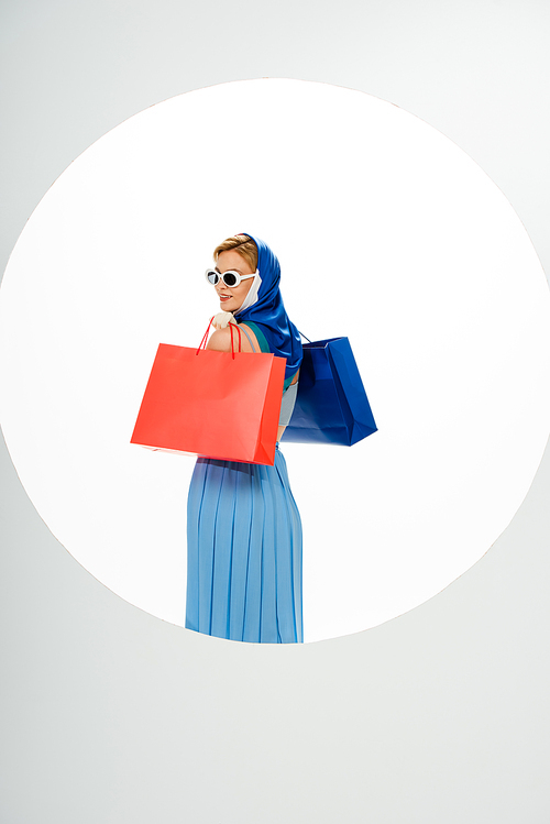 Back view of smiling woman in headscarf and sunglasses holding red and blue shopping bags behind circle on white background