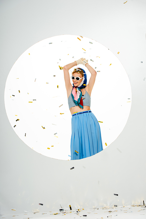 Smiling woman in headscarf and sunglasses standing under falling confetti near round hole on white background