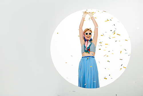 Positive girl in sunglasses standing near circle under falling confetti on white background