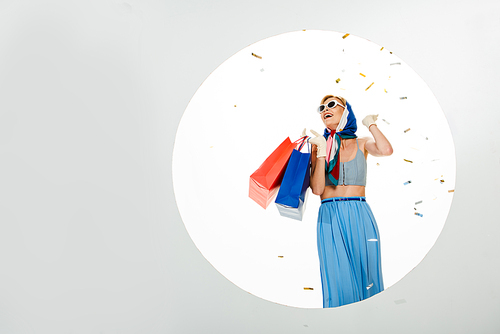 Cheerful stylish girl holding shopping bags under falling confetti near circle on white background
