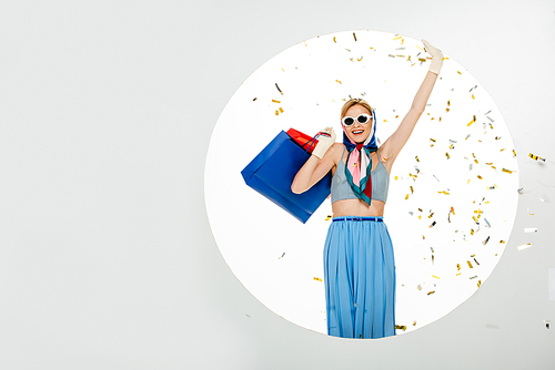 Smiling woman in sunglasses holding colorful shopping bags under falling confetti near circle on white background