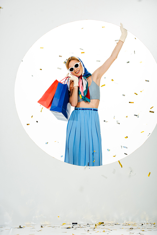 Smiling woman in headscarf and sunglasses holding shopping bags under falling confetti near circle on white background
