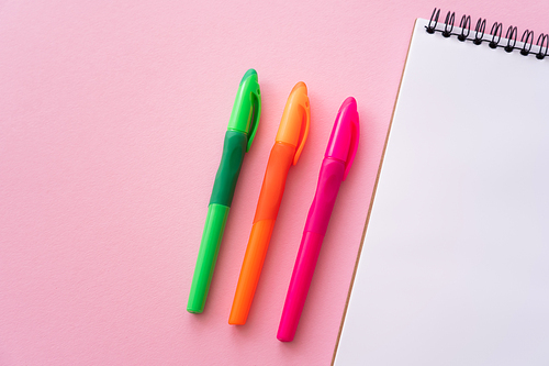 top view of colorful and bright pens near blank notebook on pink