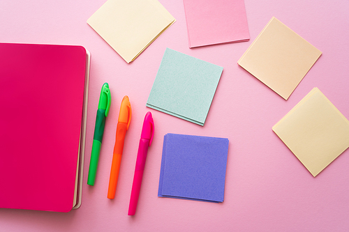 top view of colorful pens near bright notebook and blank paper notes on pink