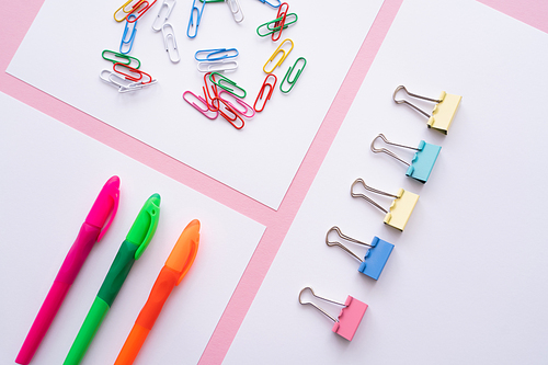 flat lay of colorful fold back clips, paper clips and pens on white papers on pink