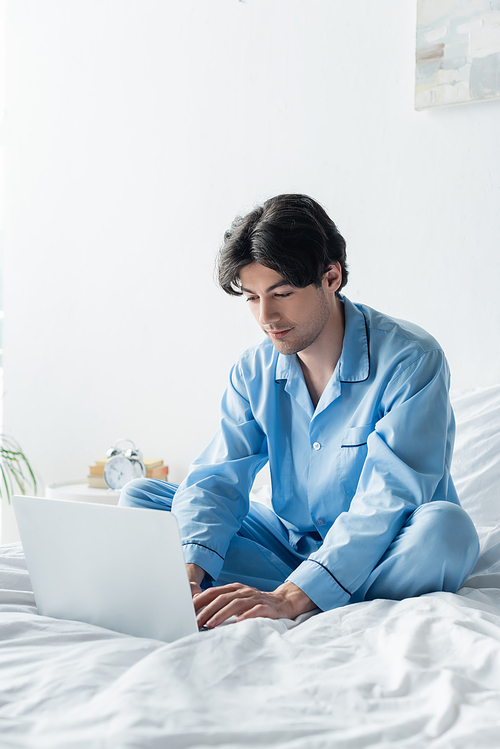 man in blue pajamas sitting on bed with crossed legs and typing on laptop