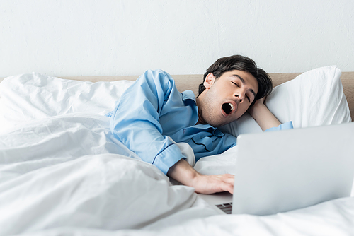 sleepy man yawning near laptop while lying in bed in morning