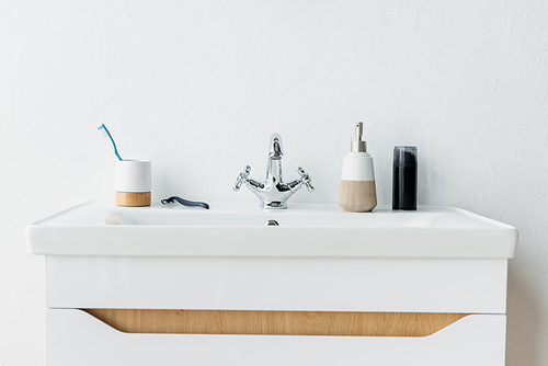 white sink with faucet, dispenser with liquid soap, shaving foam, safety razor and toothbrush