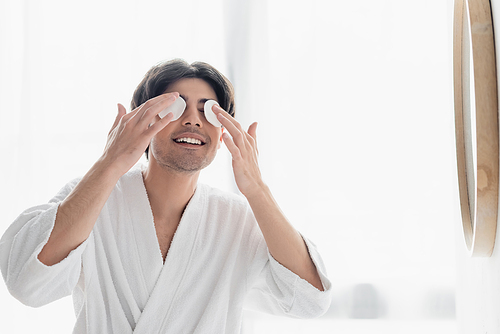 cheerful man in white bathrobe wiping eyes with cotton pads in bathroom