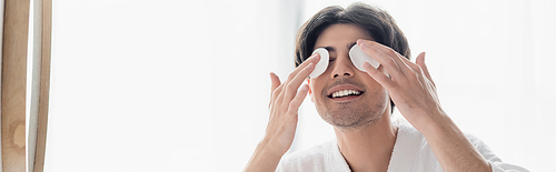 smiling brunette man wiping eyes with cotton pads in bathroom, banner