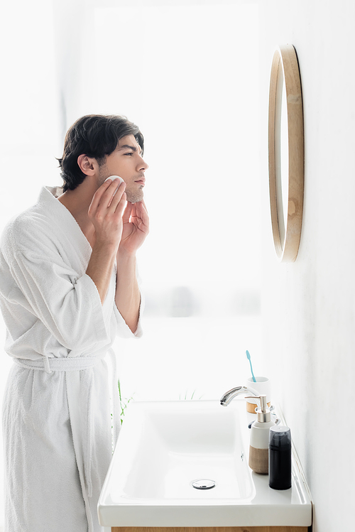 side view of man in white bathrobe cleaning face with cotton pad in bathroom